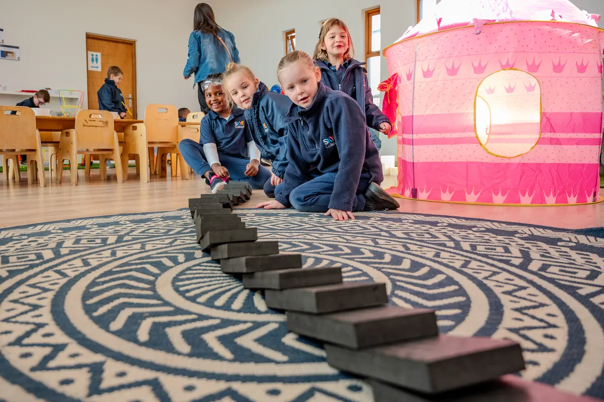 Children setting up a long domino run across a patterned classroom rug