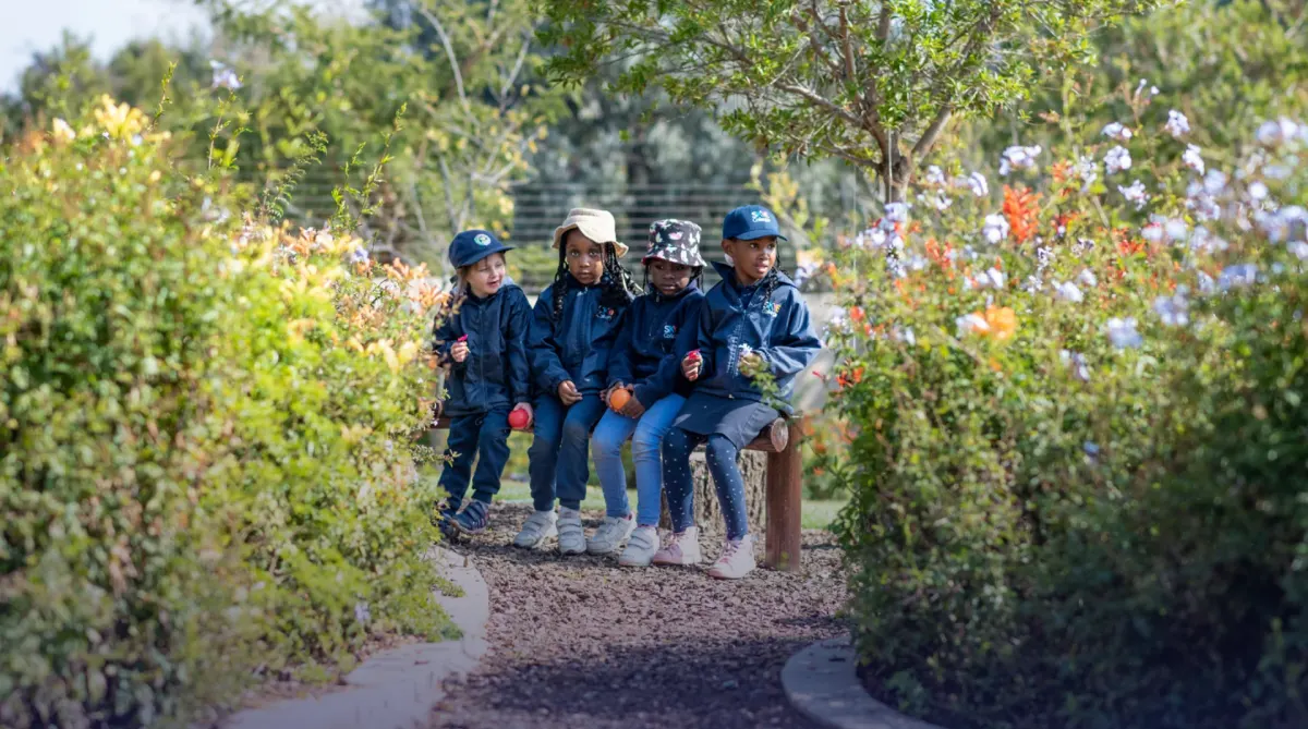 Young children in hats sitting together on a bench in a flowering garden